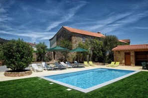 Una piscina con tumbonas y sombrillas, rodeada de un césped cuidado y un edificio de piedra con tejado de baldosas.