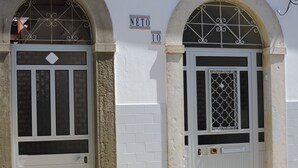 Two white doors with glass panels and decorative metal grilles, set in a building with white brick walls and a sign reading "NETO 10". - House / Villa - Fuseta (Fuseta)