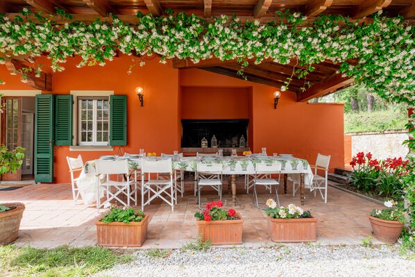 Ein Essbereich im Freien mit einem für sechs Personen gedeckten Tisch, umgeben von Stühlen, unter einer hölzernen Pergola mit Kletterpflanzen.