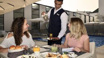 Una mesa de comedor con alimentos para el desayuno, un camarero sirviendo una bebida y una piscina al fondo.