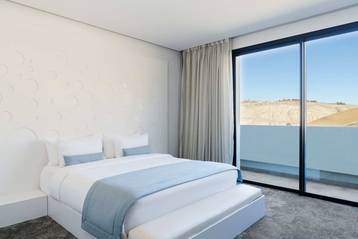 A modern hotel room with a large bed, a view of a desert landscape through floor-to-ceiling windows, and a minimalist design.