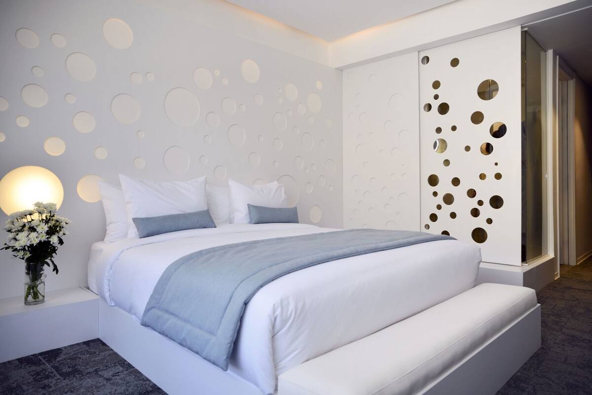 A modern bedroom with a large bed, a nightstand with a vase of flowers, and a wall with circular cut-outs.