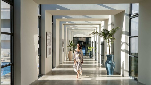 Un couloir d’hôtel moderne avec un mur en verre, un grand vase bleu et une piscine visible à l’extérieur.