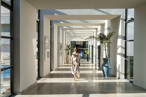 Un couloir d’hôtel moderne avec un mur en verre, un grand vase bleu et une piscine visible à l’extérieur.