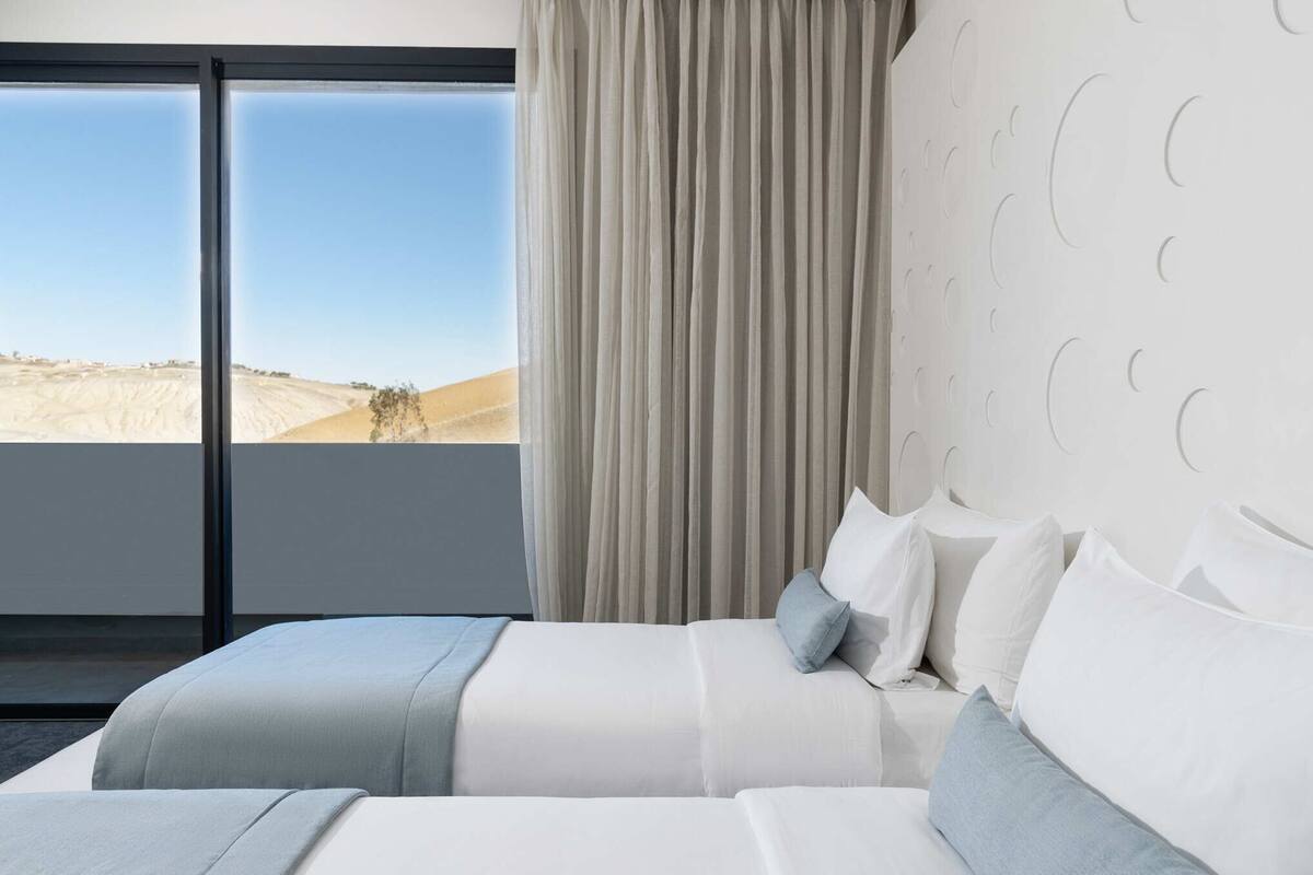 A modern hotel room with a large window, a bed with white linens, and a view of a desert landscape.