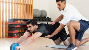 Two individuals performing an exercise with a wheel on a mat in a gym with exercise equipment in the background.