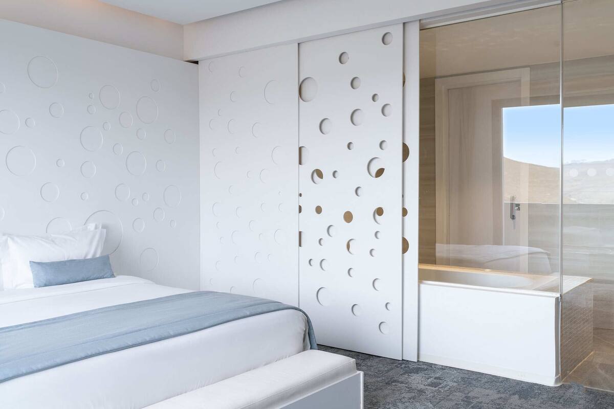 A modern bedroom with a bed, a glass shower enclosure, and a wall with circular cutouts.