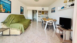 Een woonkamer met een groene bank, een eettafel met stoelen, een televisie en een keuken op de achtergrond.