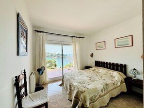 Un dormitorio con cama, una silla, una mesita de noche, lámpara, ventilador, ventana con cortinas y vista al mar.