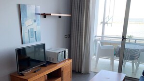 Una sala moderna con una televisión de pantalla plana, un microondas y un mueble de madera. Hay una puerta corrediza de vidrio que da a un balcón con vista al mar.