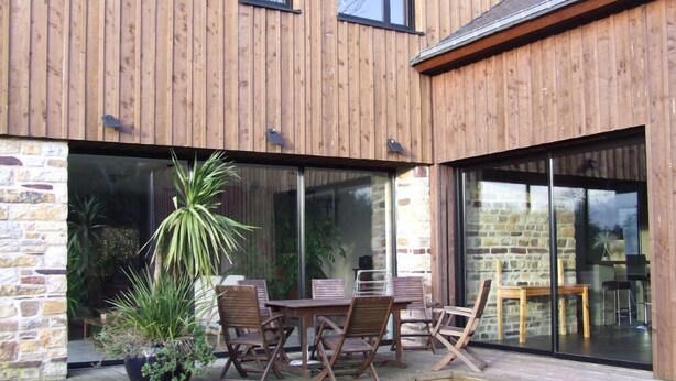 Ein modernes Haus mit Holzfassade, einer Terrasse mit Tisch und Stühlen sowie einer Steinmauer.