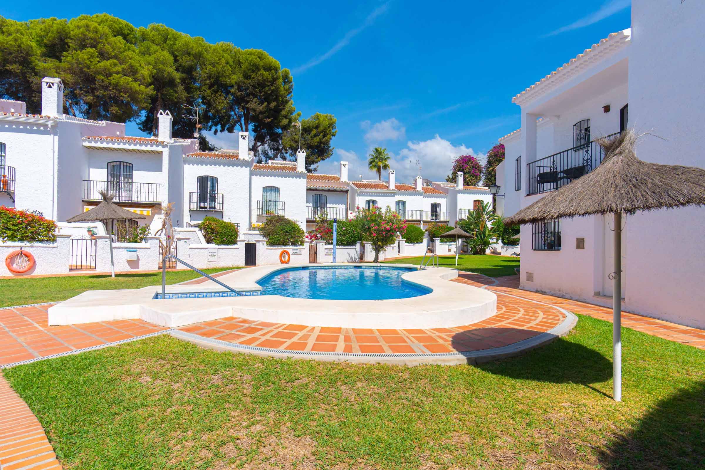 A pool area with a circular design, surrounded by white buildings with red-tiled roofs, and a thatched umbrella.