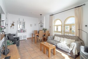 Sala de estar com sofá, mesa de centro e área de jantar com cadeiras e mesa. Há uma estante de livros e uma janela com cortinas.