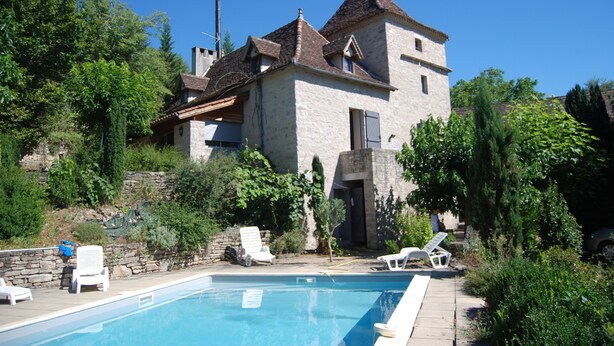 A stone house with a swimming pool, surrounded by greenery and two white lounge chairs.