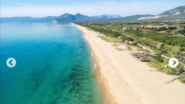 Eine Küstenlandschaft mit Sandstrand, klarem türkisblauem Wasser und Bergen in der Ferne.