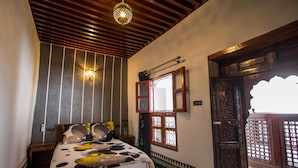 A bedroom with a wooden bed, a nightstand, a wall-mounted light, a window with a decorative frame, and a patterned floor. - Dar el Mathaf (Fes)