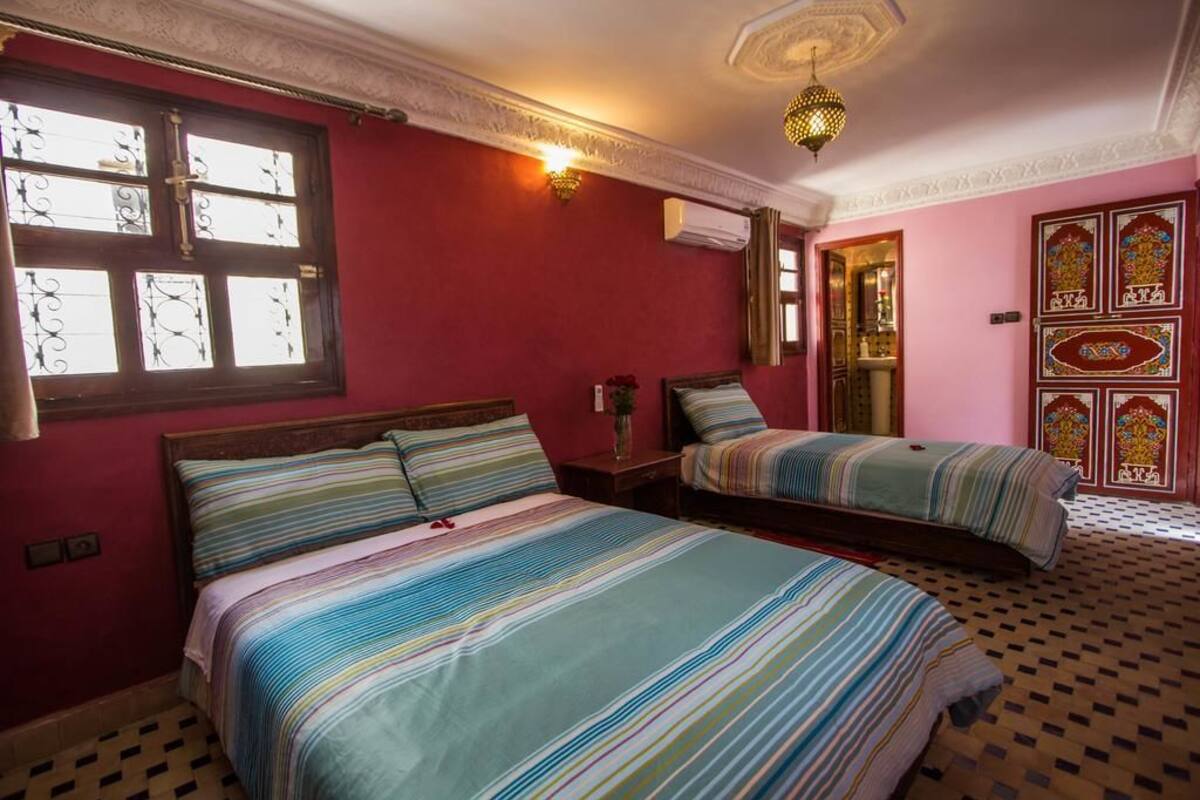 Una habitación con dos camas, una ventana con rejas decorativas, aire acondicionado de pared y piso estampado.