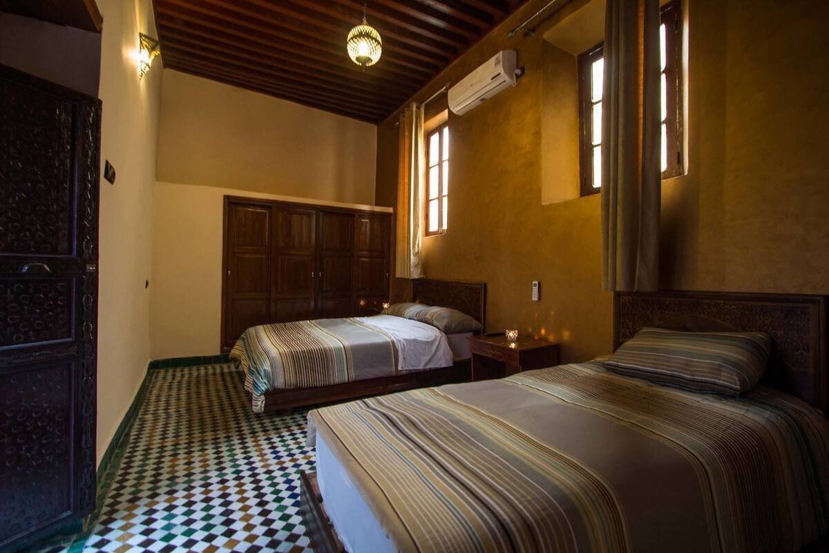 Una habitación de hotel con dos camas, piso estampado, vigas de madera en el techo y una ventana con cortinas.