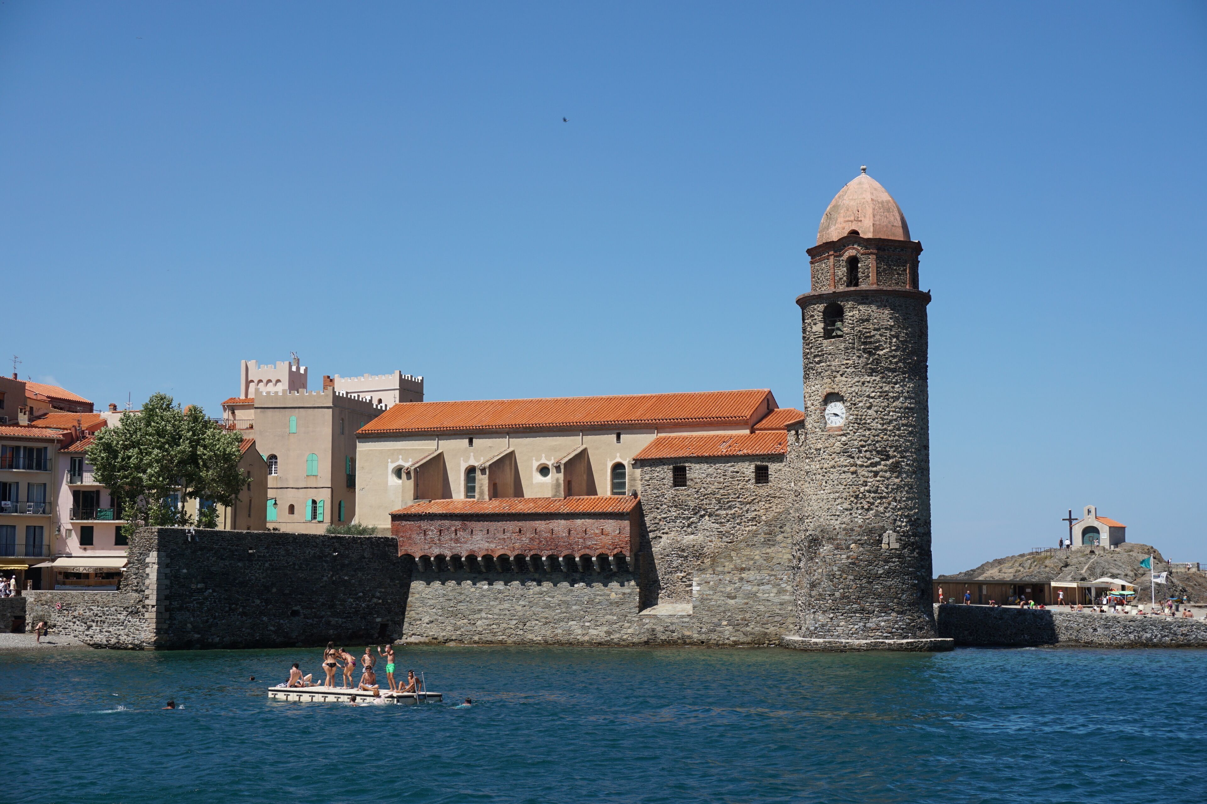Una torre de pedra històrica amb un rellotge, una barca petita amb gent i edificis amb teulades vermelles.