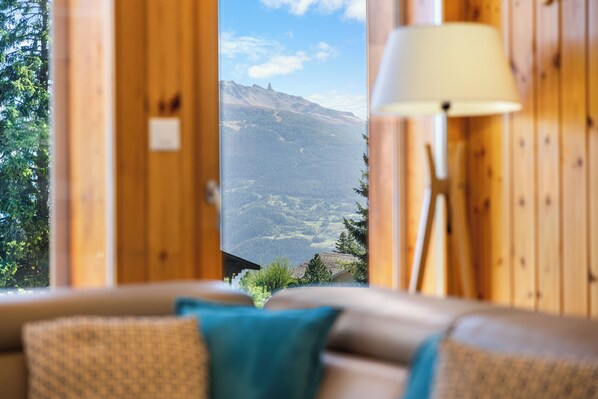 Una habitación acogedora con detalles de madera, vista a un paisaje montañoso a través de una ventana, y una lámpara a la derecha.