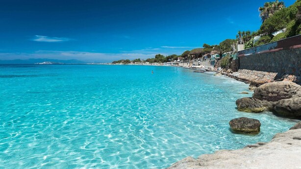 Una playa costera con agua turquesa cristalina, costa rocosa y edificios a lo largo de la orilla.
