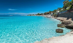 A coastal beach with clear turquoise water, rocky shoreline, and buildings along the edge.