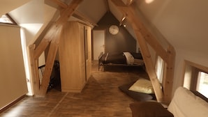 A loft bedroom with exposed wooden beams, a bed with a headboard, a clock on the wall, and a window providing natural light.