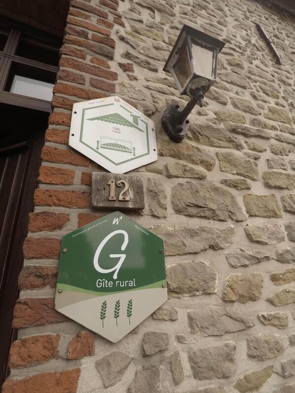 A stone wall with three signs: a rural Gîte sign, a number 12 sign, and a green sign with a white 'G' and wheat stalks.