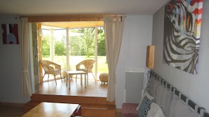 A living room with a sofa, a wooden table, and two wicker chairs. There is a sliding glass door leading to an outdoor area with a lawn and trees.