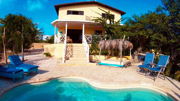 A two-story building with a pool, blue lounge chairs, and a thatched-roof structure. - Beautiful detached villa with a view over Klein Bonaire. (Santa Barbara, Kralendijk)