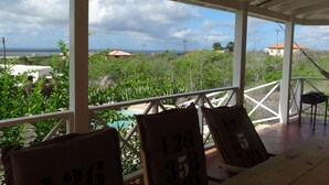 A covered outdoor seating area with chairs numbered 126, 158, and 35, overlooking a lush landscape with a view of the ocean and distant buildings. - Beautiful detached villa with a view over Klein Bonaire. (Santa Barbara, Kralendijk)