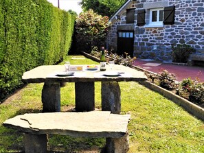 Une table en pierre, avec une bouteille, des verres et une assiette de nourriture, se trouve dans un coin d’un jardin, avec un bâtiment en pierre en arrière-plan.