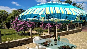 Un espace extérieur aménagé pour prendre les repas, avec un sol en pierre, des chaises en plastique blanches, une table sur laquelle se trouve une bouteille de vin, et un parasol rayé qui offre de l’ombre.