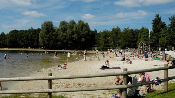 Une plage où des gens se baignent et se détendent sur le sable, une clôture en bois au premier plan et des arbres en arrière-plan.