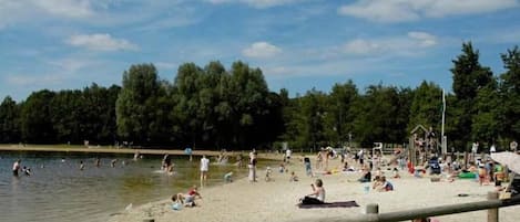 A beach with people swimming and relaxing on the sand, a wooden fence in the foreground, and trees in the background.