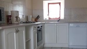 A kitchen with white cabinets, a stove, an oven, and a dishwasher. There is a window with a red frame and a small shelf with a kettle. - Le Cosquer farm (Saint-Connec)