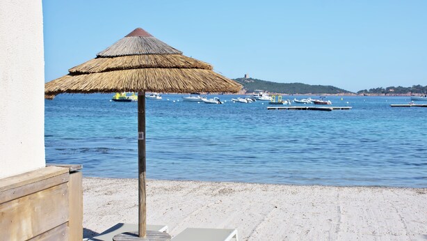 Une plage avec un parasol en chaume, des chaises longues et des bateaux sur l’eau.
