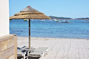 A beach with a thatched umbrella, lounge chairs, and boats in the water.