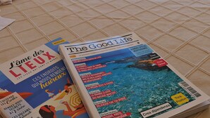 Une chambre d’hôtel avec un lit, une table de chevet avec une lampe, deux chaises près d’un balcon et un magazine sur le lit.