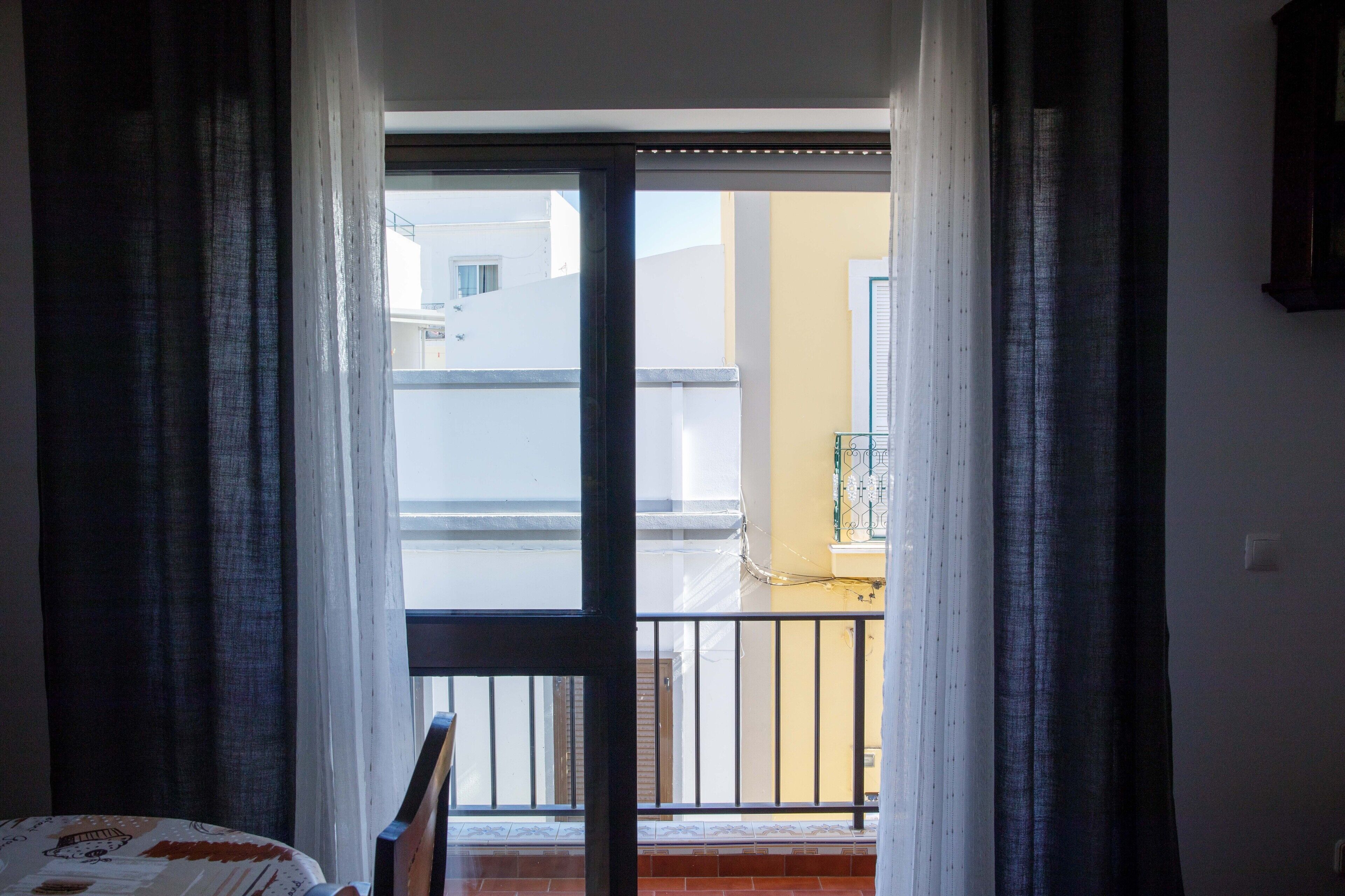 Ein Zimmer mit Bett, einem Fenster mit Vorhängen und Blick auf einen Balkon mit Geländer.