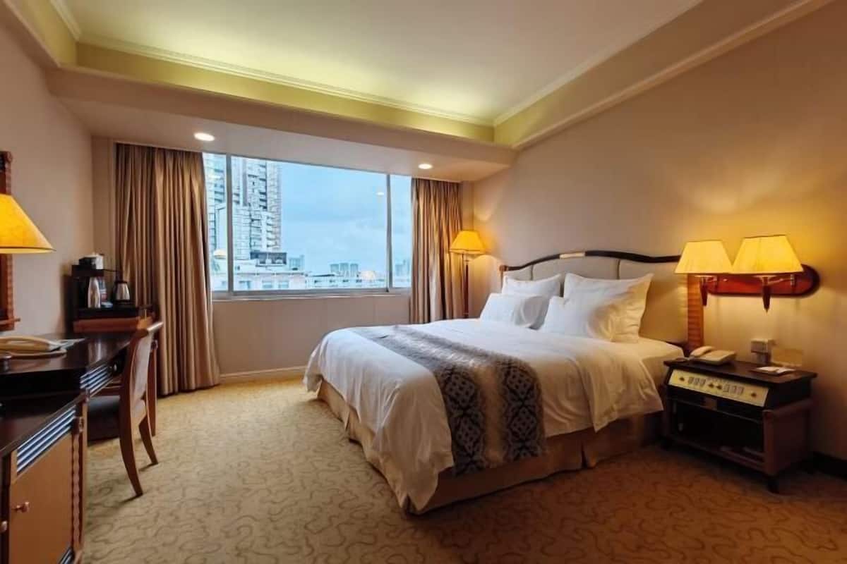 A hotel room with a large bed, a desk, a chair, a nightstand with a lamp, and a view of the city.