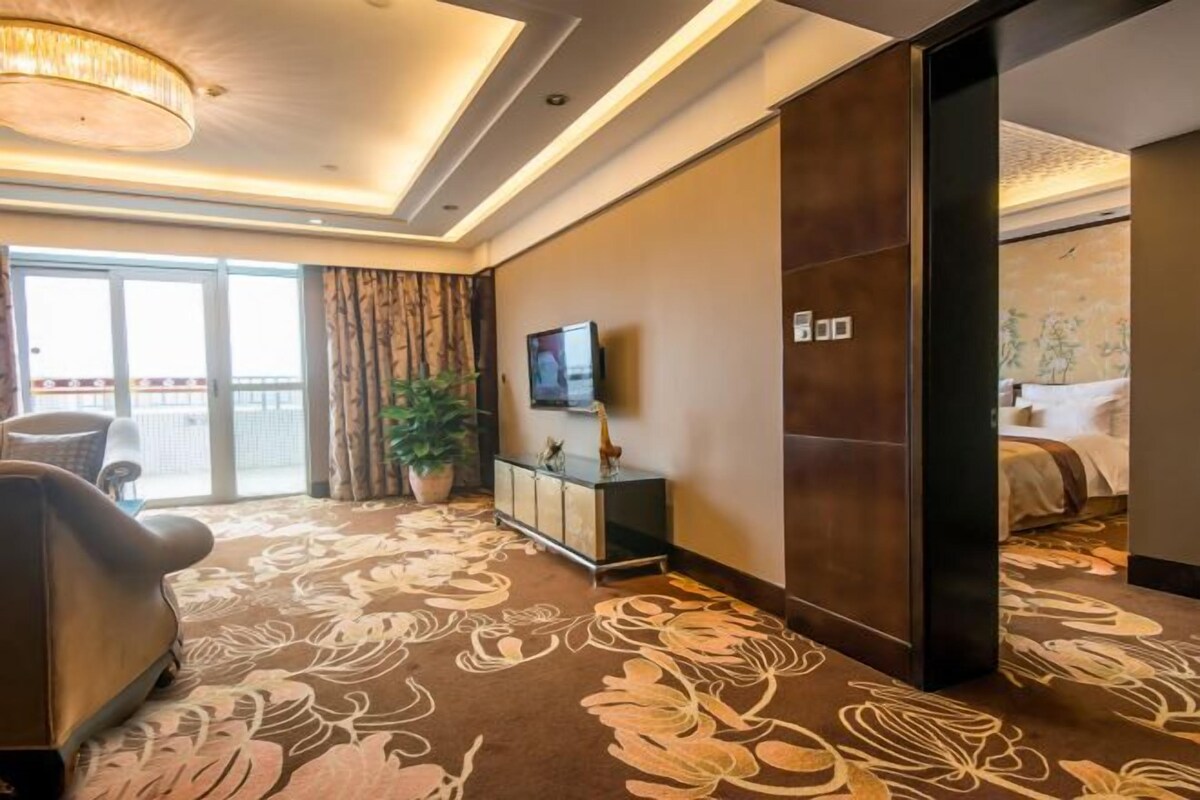 A hotel room with a large bed, a TV, a potted plant, and a view of the outside balcony.