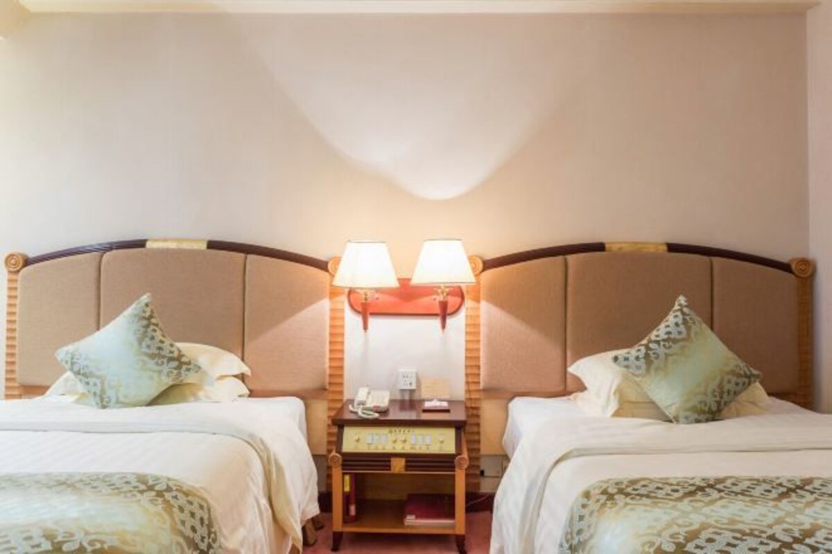 A hotel room with two beds, each with patterned bedding and pillows. There is a bedside table with a lamp between the beds.