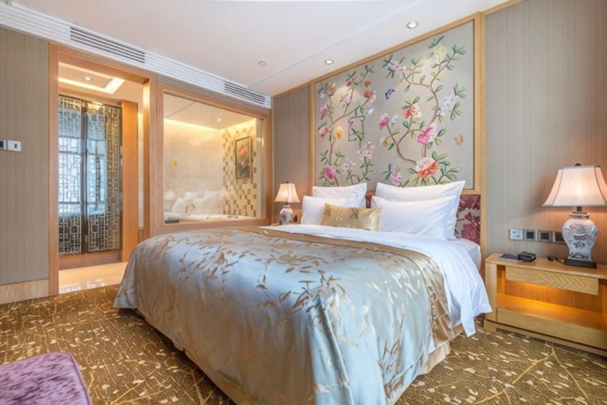 A hotel room with a large bed, a nightstand with a lamp, a floral wall mural, and a sliding door leading to a bathroom.