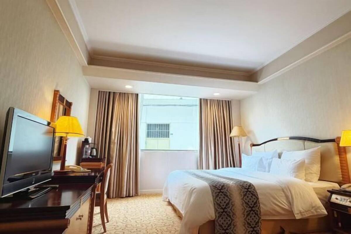 A hotel room with a bed, a desk with a television, a chair, and a window with curtains.