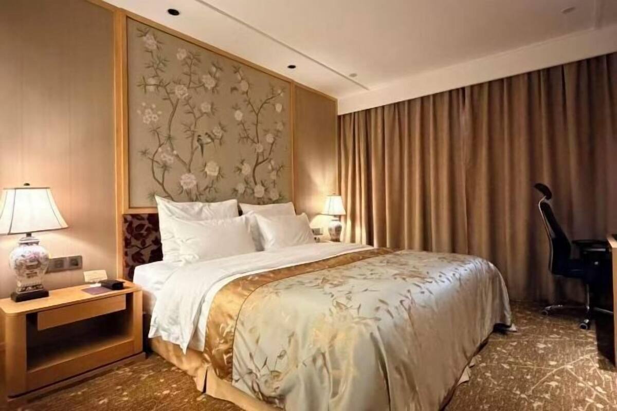 A hotel room with a large bed, a nightstand with a lamp, a desk with a chair, and a decorative wall panel.