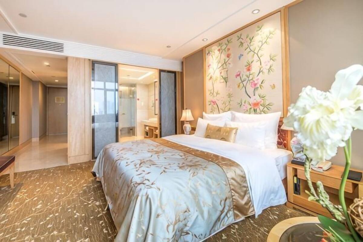 A hotel room with a large bed, a bedside table with a lamp, a decorative wall, and a view of the hallway.