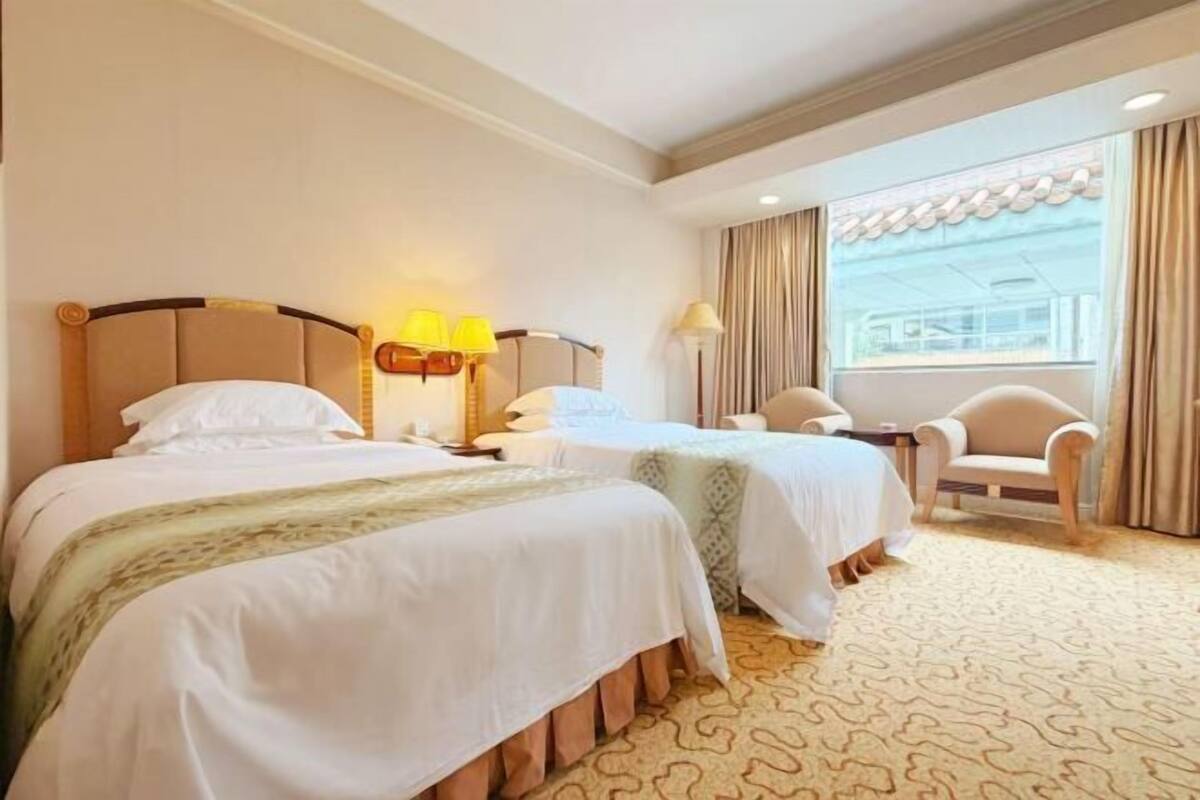 A hotel room with two beds, a chair, a window with curtains, and a lamp.