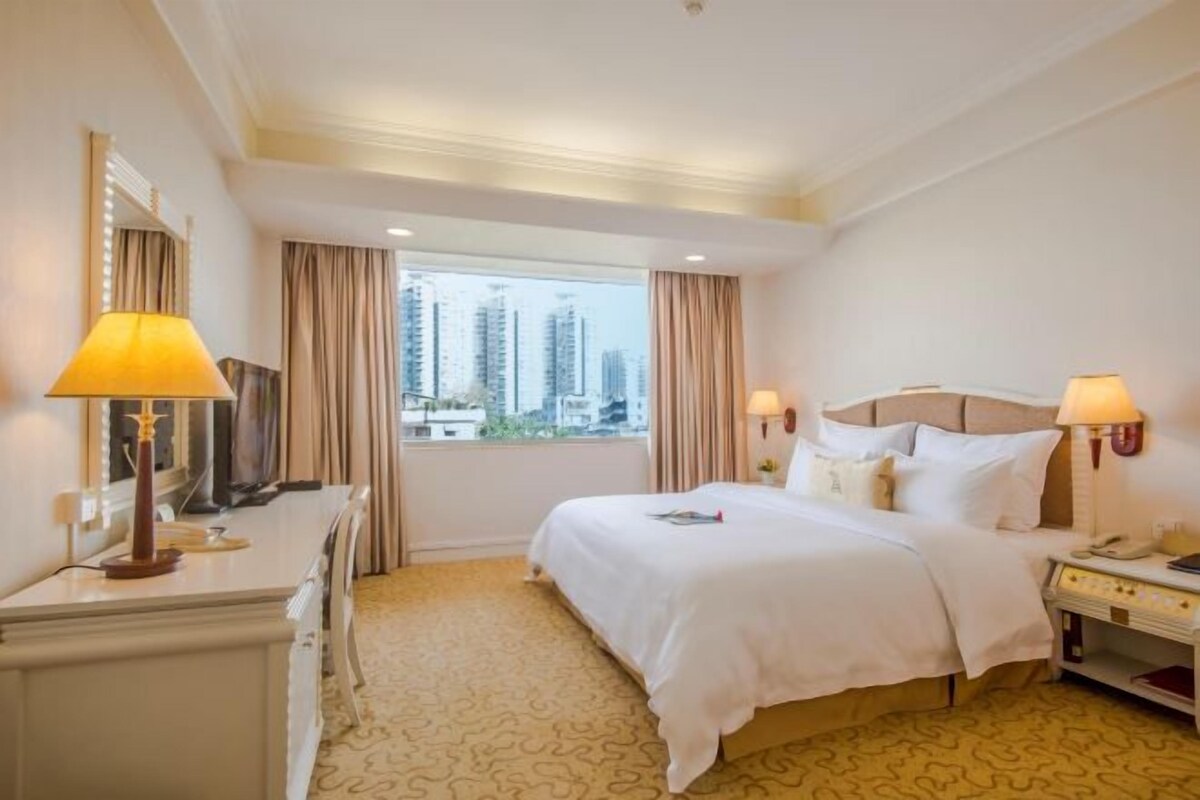 Une chambre d’hôtel avec un grand lit, deux lampes de chevet, un bureau avec une lampe, une commode et une grande fenêtre avec des rideaux.