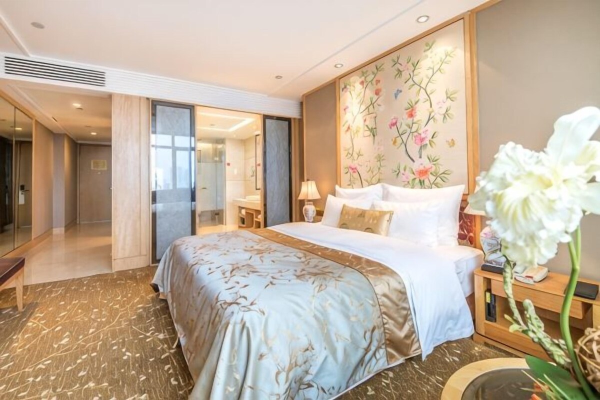A hotel room with a large bed, a bedside table with a lamp, a decorative wall, and a view of the hallway.